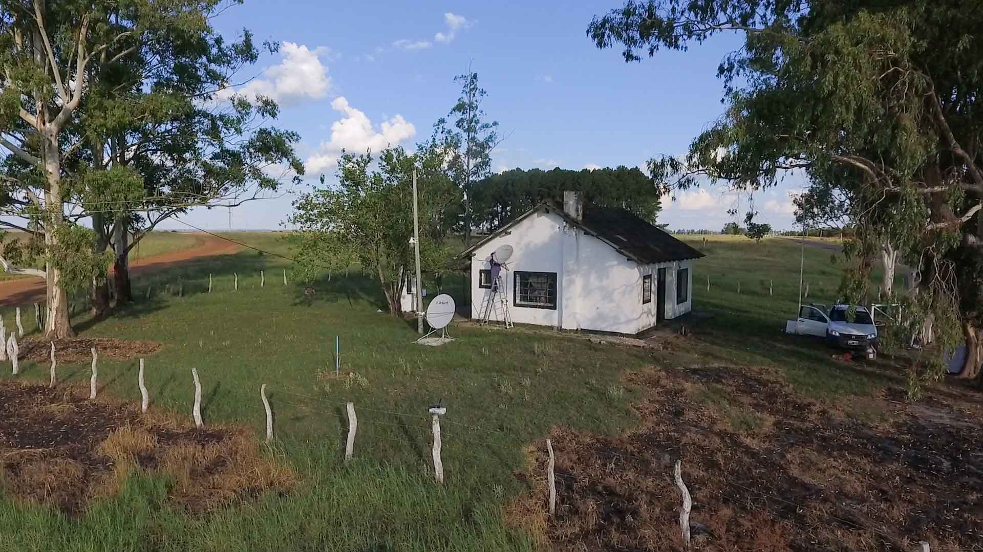 Desde un dron, una escuela rural rodeada de un campo verde. En el piso hay instalada una antena de internet y en la pared una de Televisión digital.